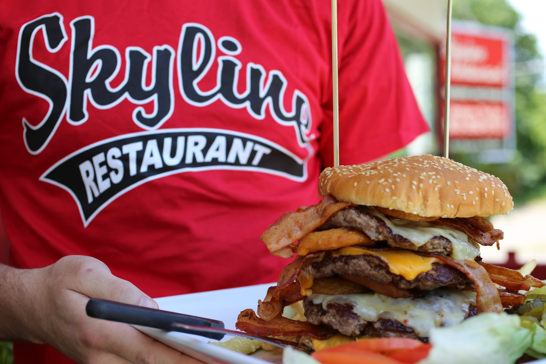skyline restaurant tshirt holding a 4-patty burger with layers of swiss and american cheese and bacon held together with wooden sticks
